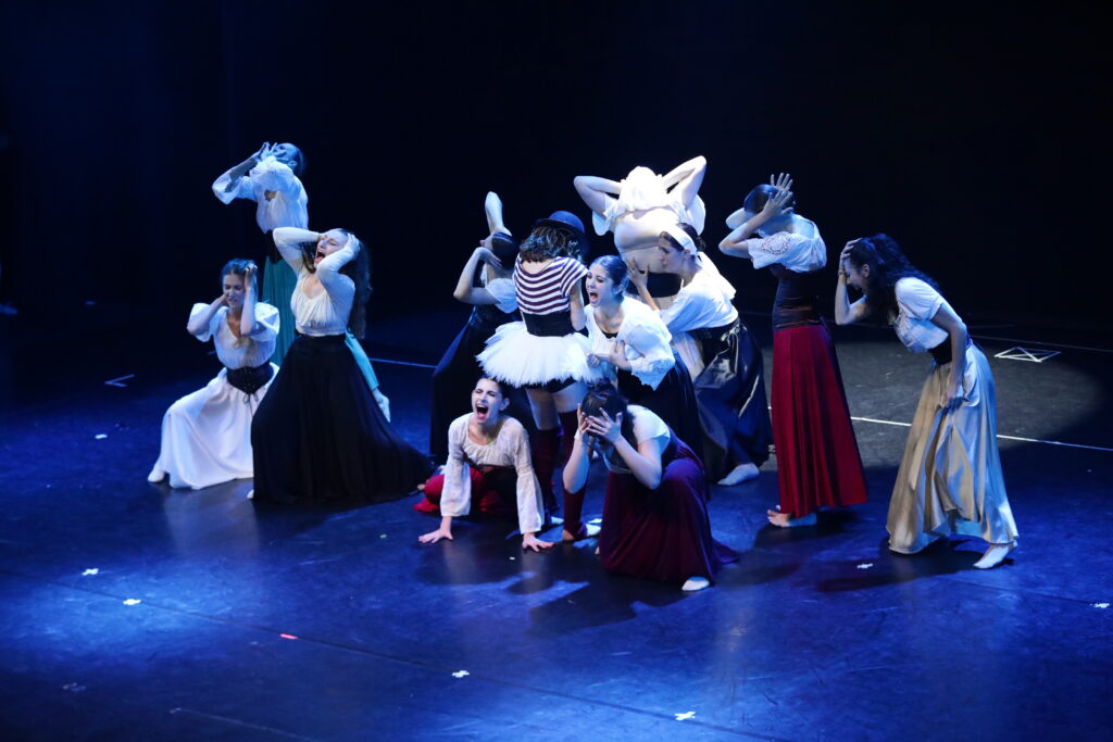 Gruppo di ballerini in abiti storici, esprimono emozioni intense su un palco illuminato.