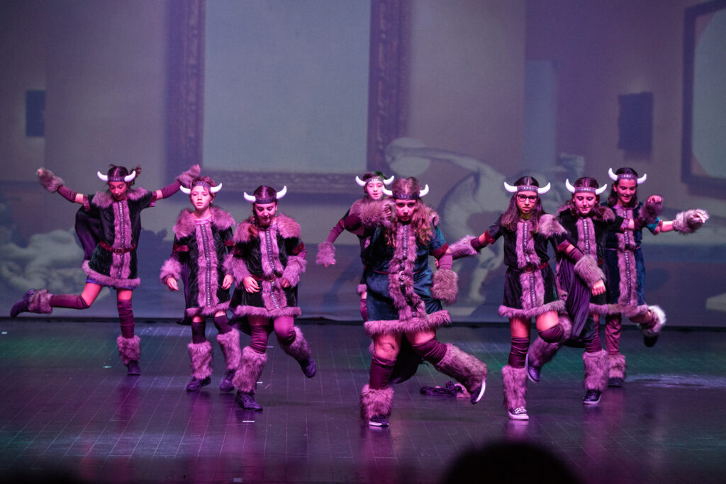 Gruppo di danzatori in costumi vichinghi, eseguono una coreografia energica su un palco illuminato.