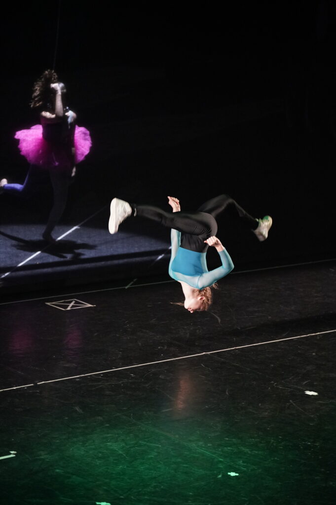 Danzatore in volo durante una performance, riflesso sullo sfondo, indossa un costume colorato.