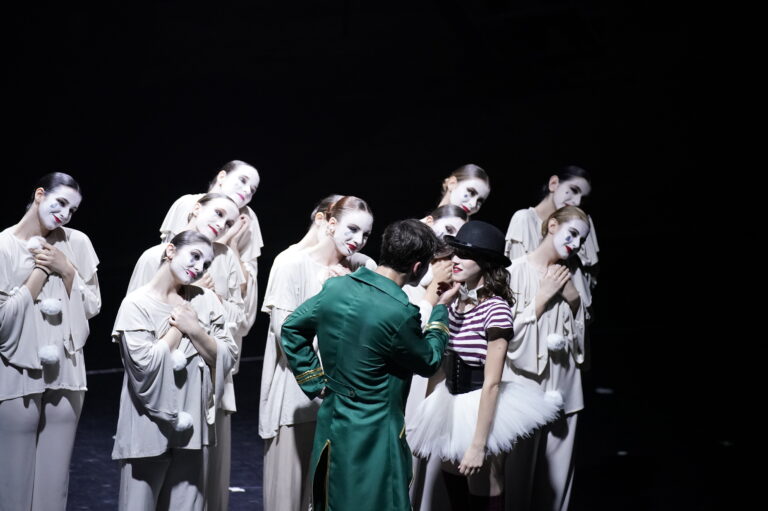 Gruppo di ballerini in abiti chiari e trucco bianco, mentre due danzatori si avvicinano al centro del palco.