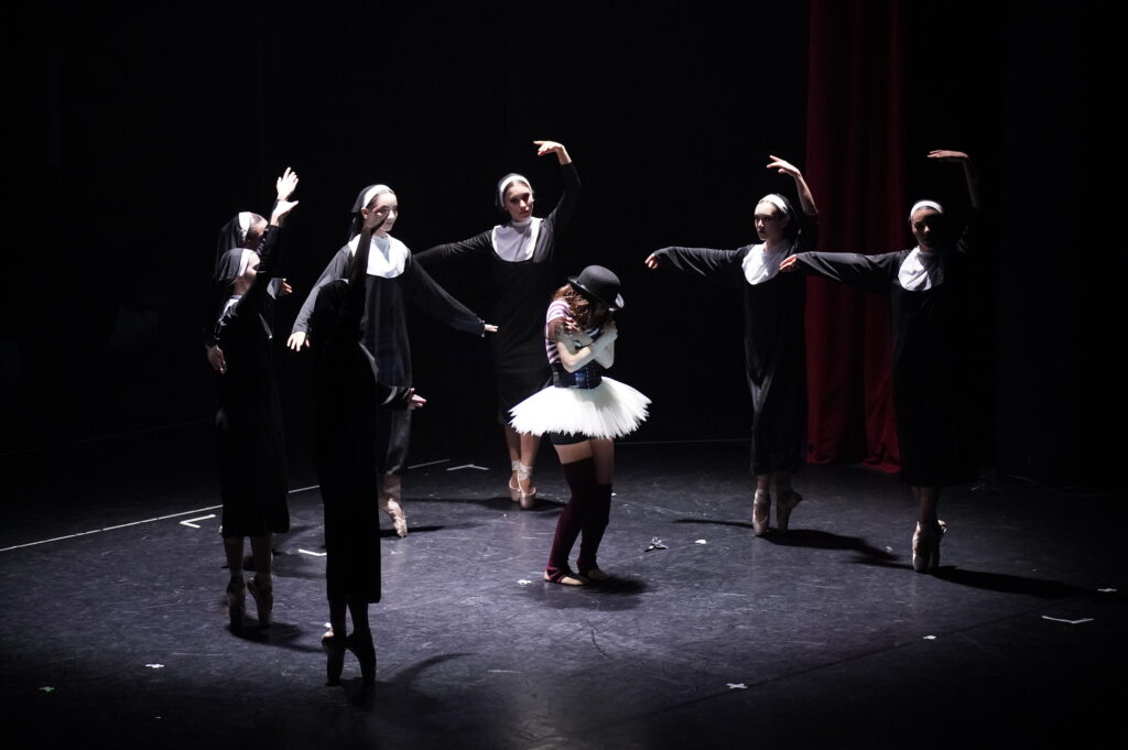 Un gruppo di ballerine in abiti scuri circonda una danzatrice con tutù bianco, in un momento di danza teatrale.