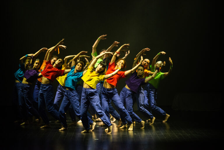 Gruppo di ballerini in magliette colorate e pantaloni blu, in una coreografia dinamica su un palco illuminato.