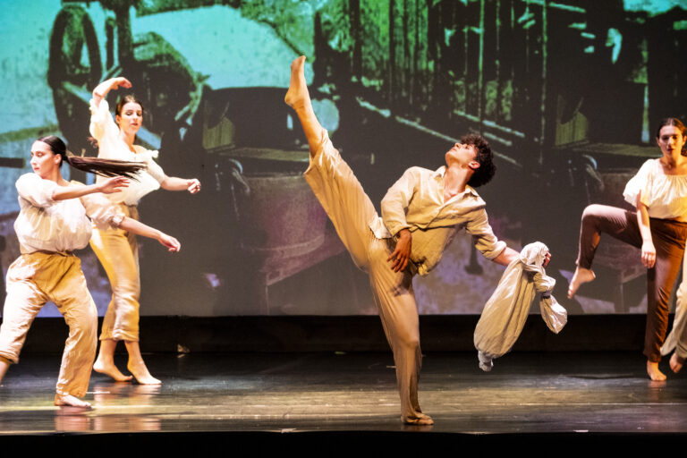 Gruppo di ballerini in scena, uno esegue un alto movimento di danza mentre gli altri lo osservano.