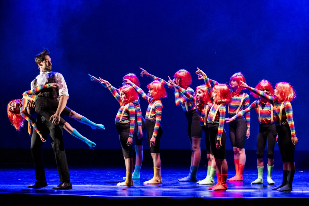 Gruppo di ballerini con parrucche arancioni e costumi colorati, in scena durante una performance teatrale.