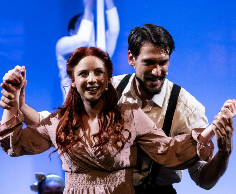 Due persone sul palco si tengono per mano, sorridendo, in un momento di danza e celebrazione.