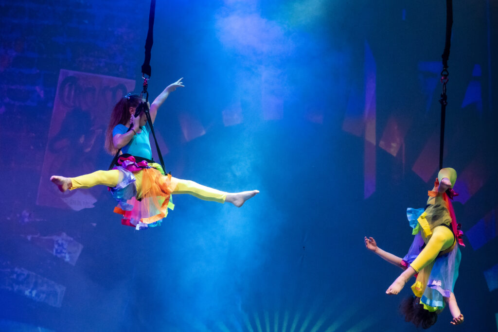 Due artisti acrobatici sospesi in aria, indossano costumi colorati e si esibiscono in un numero di danza.