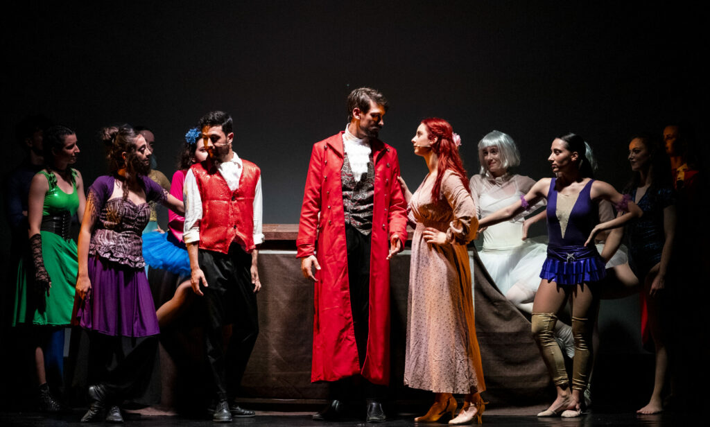 Gruppo di performer in abiti teatrali, con scene di interazione e danza sul palco.