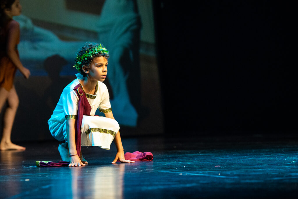 Bambino in costume teatrale, in posa dinamica su un palco, con una corona di foglie e un drappo colorato.
