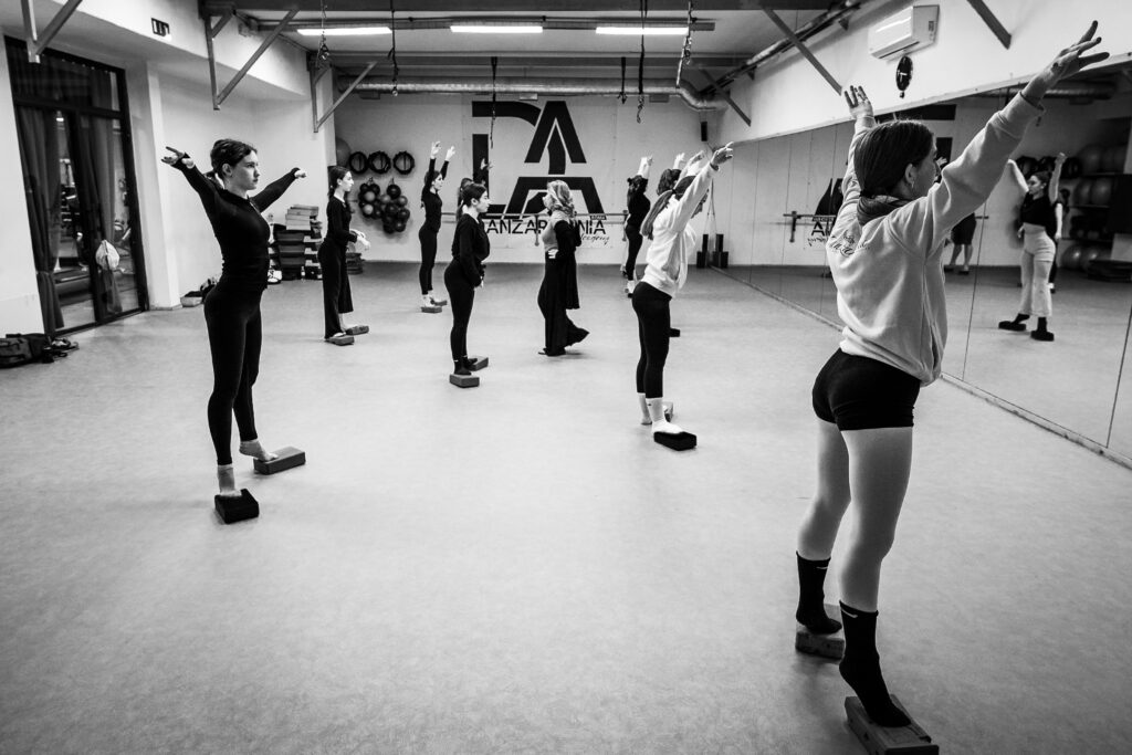 Gruppo di ballerine in sala danza, in posizione di stretching con le braccia sollevate.