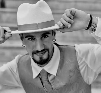 Uomo con cappello bianco e cravatta, sorride mentre regge il cappello con entrambe le mani.