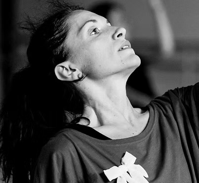 Donna con capelli scuri e lunghi, in posa di danza, guarda verso l'alto con espressione concentrata.