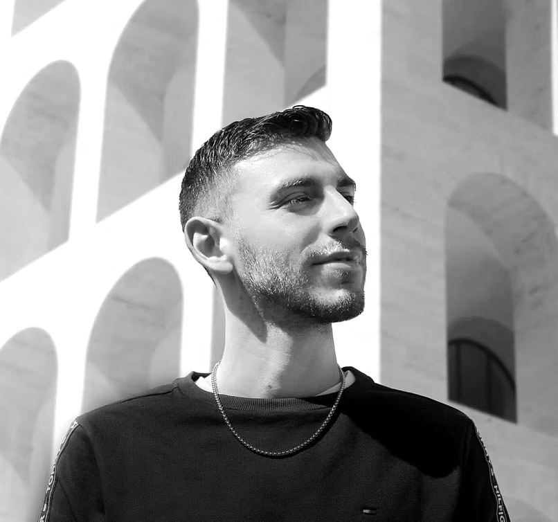 Giovane con barba e capelli corti, in primo piano, davanti a un'architettura moderna in bianco e nero.