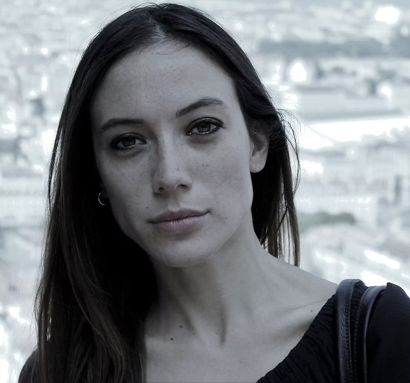 Donna con capelli lunghi e scuri, sguardo intenso, sfondo urbano sfocato.