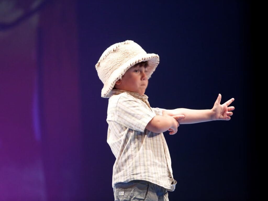Bambino con cappello di paglia, in posa su un palco, mentre allunga un braccio verso il pubblico.