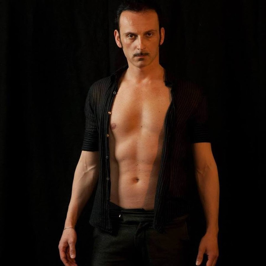 Uomo con torso scoperto, indossa una camicia nera aperta e pantaloni scuri, in piedi su sfondo nero.