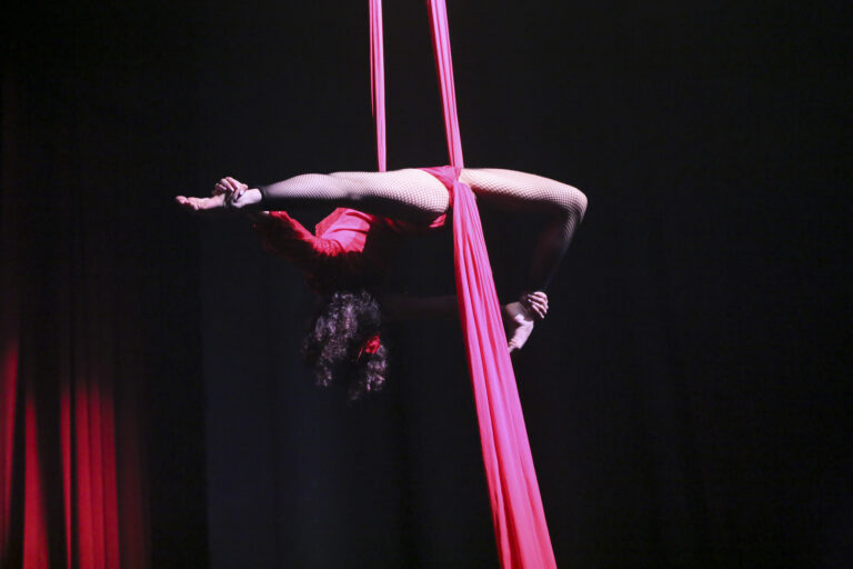 Artista acrobatica sospesa in un drappo rosso, in una posa elegante e dinamica, sullo sfondo scuro.