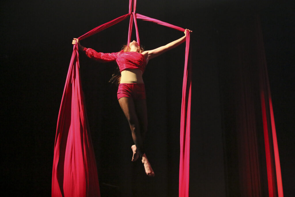 Artista di circo sospesa in aria con tessuti rossi, in un momento di danza acrobatica.