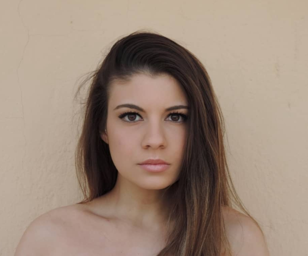 Ritratto di una giovane donna con capelli lunghi e lisci, sguardo intenso e sfondo neutro.