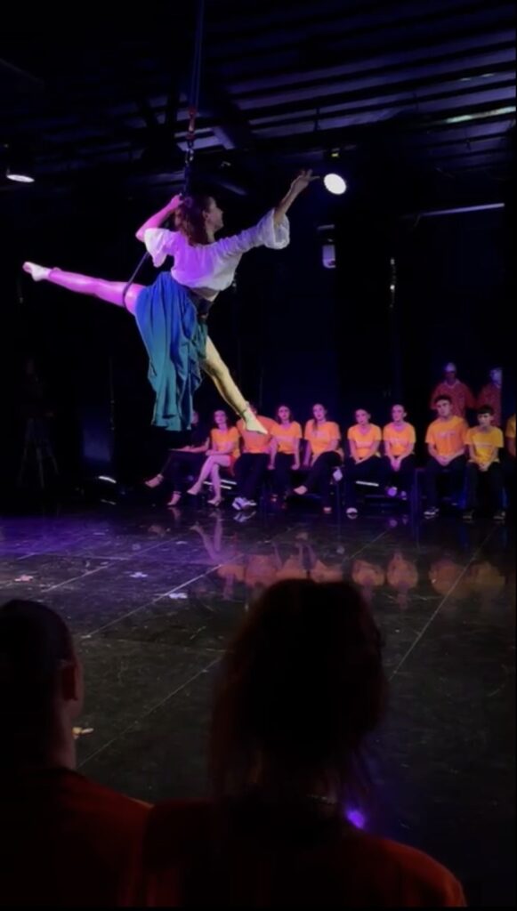 Danza aerea con una performer in volo, mentre il pubblico osserva in abiti coordinati.